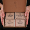 A gift set of soaps from Three Hills Soap displayed in a brown cardboard box. The set includes four bars of soap labeled "Lavender Rosemary," "Ylang Ylang Patchouli," "Bergamot Verbena," and "Geranium Palmarosa." The box is open to show the soaps, which are neatly arranged in two rows of two. The company's logo is printed on the bottom right corner of the box lid.