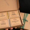 A gift set of soaps from Three Hills Soap displayed in a brown cardboard box. The set includes four bars of soap labeled "Carrot," "Pure Calendula," "Dead Sea Mud," and "Chamomile Calendula with Oats." The box is open to show the soaps, which are neatly arranged in two rows of two. The box is decorated with a green ribbon tied in a simple knot on the right side. The company's logo is printed on the bottom right corner of the box lid.