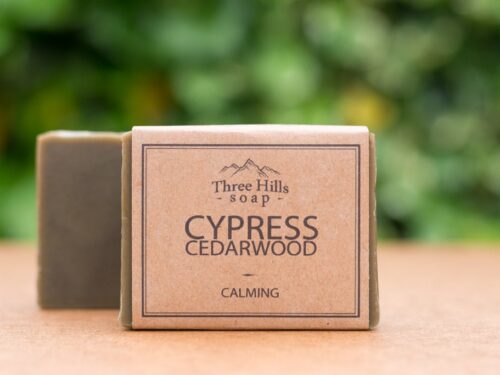Image of a Three Hills Soap bar in Cypress Cedarwood scent. The soap is labeled 'Calming' and comes in a natural, eco-friendly package. The earthy-toned soap bar is positioned in the foreground with a blurred, green background.