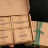 A gift set of soaps from Three Hills Soap displayed in a brown cardboard box. The set includes four bars of soap labeled "Sweet Orange Spearmint," "Lemongrass Rosemary," "Ginger Lime," and "Lemon Mandarin." The box is open to show the soaps, which are neatly arranged in two rows of two. The box is decorated with a green ribbon tied in a simple knot on the right side. The company's logo is printed on the bottom right corner of the box lid.