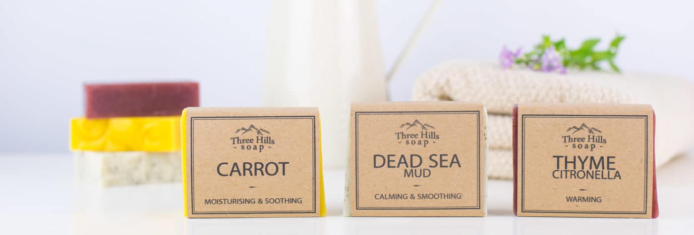 A collection of handmade soap bars in various colors and shapes, neatly arranged on a white surface. Some bars feature natural elements like dried lavender, oats, and decorative herbs, highlighting the use of organic and natural ingredients.