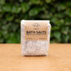 The image shows a package of bath salts placed on a wooden surface with a green leafy background. The packaging is transparent, revealing a coarse, speckled mixture inside, labeled with a brown paper label. The label reads "Three Hills Soap" at the top, followed by "EXOTIC BATH SALTS" in large, bold letters. Below, it states "HIBISCUS & YLANG YLANG" in smaller text. Additional details at the bottom of the label indicate that the product is "100% NATURAL" and "VEGAN FRIENDLY."