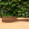 Biodegradable and eco-friendly Liquid Wood Comb, made from renewable materials, placed on a wooden surface with a green leafy background. The comb features a smooth handle and fine teeth, perfect for sustainable hair care.