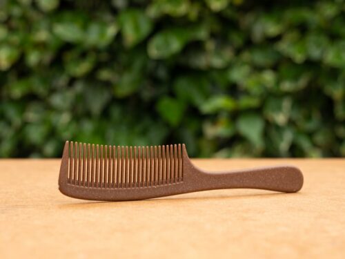 Biodegradable and eco-friendly Liquid Wood Comb, made from renewable materials, placed on a wooden surface with a green leafy background. The comb features a smooth handle and fine teeth, perfect for sustainable hair care.