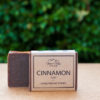 A bar of soap labeled "Three Hills Soap - Cinnamon Soap - Gingerbread Spiced" is shown in the image. The soap is a rich brown color and partially wrapped in a simple, rustic brown paper label with black text. The background is blurred, featuring green foliage, suggesting the photo was taken outdoors. The soap stands on a flat, light-colored surface. The overall presentation highlights the product's warm and spiced qualities, reminiscent of gingerbread.