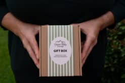 The image shows a pair of hands holding a closed gift box, wrapped in kraft paper with a simple, natural string tied in a bow. The box is plain and minimalistic, reflecting an eco-friendly and artisanal approach. The hands gently cradle the box, suggesting care and the anticipation of giving or receiving a thoughtful, handcrafted gift. The overall feel of the image emphasizes simplicity, natural beauty, and the quality of the contents within.