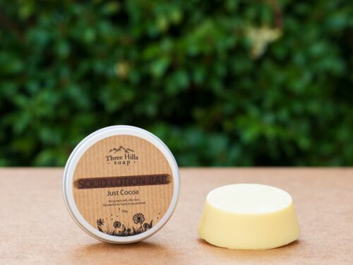 Just Cocoa solid lotion bar and labeled tin on flat surface with green blurred background