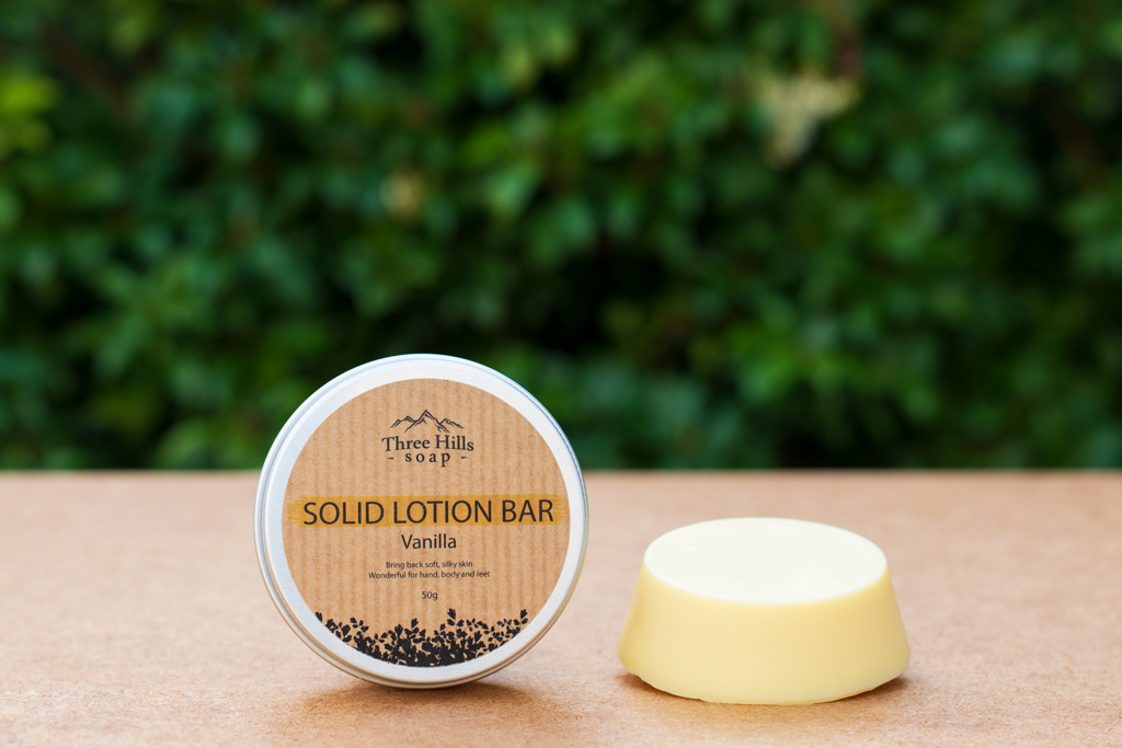 A tin container and a solid lotion bar are displayed on a flat surface against a blurred green foliage background. The tin container is labeled "Three Hills Soap - Solid Lotion Bar - Vanilla" and includes details about its benefits for bringing back soft, silky skin and being suitable for hands, body, and feet. The solid lotion bar, positioned next to the container, is a smooth, creamy yellow disc. The clean and natural presentation emphasizes the product's nourishing and eco-friendly qualities, ideal for an e-commerce listing.