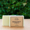 A bar of soap labeled "Three Hills Soap - Bergamot Verbena - Cheerful" is shown in the image. The soap is a light green color and partially wrapped in a simple, rustic brown paper label with black text. The background is blurred, featuring green foliage, suggesting the photo was taken outdoors. The soap stands on a flat, light-colored surface. The overall presentation highlights the product's natural and uplifting qualities.