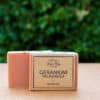 A bar of soap labeled "Three Hills Soap - Geranium Palmarosa - Romantic" is shown in the image. The soap is pinkish in color and partially wrapped in a simple, rustic brown paper label with black text. The background is blurred, featuring green foliage, indicating that the photo was taken outdoors. The soap stands on a flat, light-colored surface.