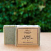 A bar of soap labeled "Three Hills Soap - Lime Patchouli - Fresh & Earthy" is shown in the image. The soap is a light green color with small speckles and is partially wrapped in a simple, rustic brown paper label with black text. The background is blurred, featuring green foliage, suggesting the photo was taken outdoors. The soap stands on a flat, light-colored surface. The overall presentation emphasizes the product's natural and refreshing qualities.