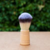 The image shows a shaving brush with a wooden handle placed on a flat surface. The bristles are dense and have a gradient from a light base to dark tips. The background is blurred, featuring green foliage, suggesting an outdoor setting.