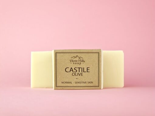 The image shows a bar of soap labeled "Three Hills Soap" with the main text on the packaging reading "CASTILE OLIVE." Below this, the description "NORMAL - SENSITIVE SKIN" is displayed. The soap has a soft, pale creamy color, typical of Castile soap made primarily from olive oil, and appears smooth in texture. It is set against a soft pink background, and the packaging is made of simple, natural-colored paper, consistent with the other soaps in this series. The overall presentation is clean and minimalistic, highlighting the gentle and nourishing qualities of the soap, especially suitable for normal and sensitive skin types.