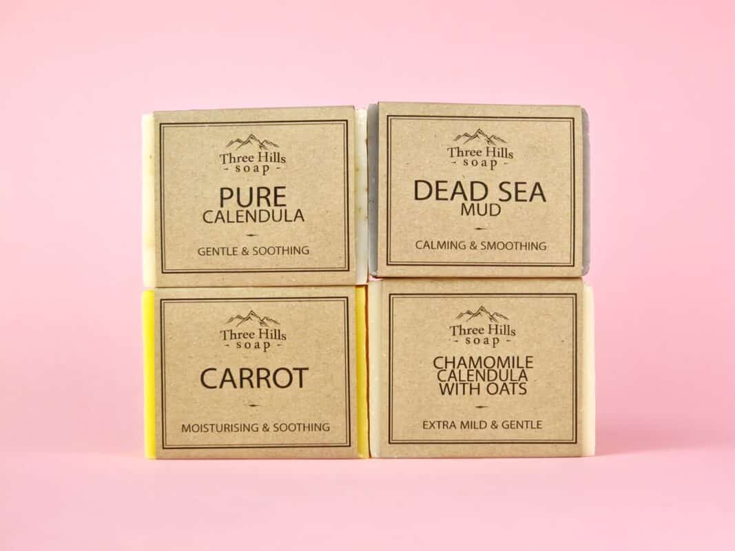 A selection of handmade soaps by Three Hills Soap, displayed on a pink background. The collection includes Pure Calendula (Gentle & Soothing), Dead Sea Mud (Calming & Smoothing), Carrot (Moisturising & Soothing), and Chamomile Calendula with Oats (Extra Mild & Gentle). Each soap bar is wrapped in minimalist packaging, highlighting its natural ingredients and intended benefits for sensitive skin.