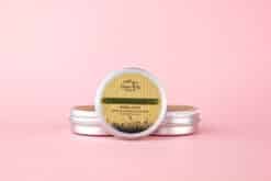 Tamanu Oil Herbal Balm with Chickweed & Plantain from Three Hills Soap, displayed in a small metal tin with a screw-top lid, placed against a pastel pink background. The label on the tin features natural design elements with earthy tones, highlighting the product’s calming and soothing properties.