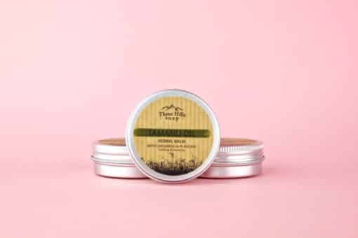 Tamanu Oil Herbal Balm with Chickweed & Plantain from Three Hills Soap, displayed in a small metal tin with a screw-top lid, placed against a pastel pink background. The label on the tin features natural design elements with earthy tones, highlighting the product’s calming and soothing properties.