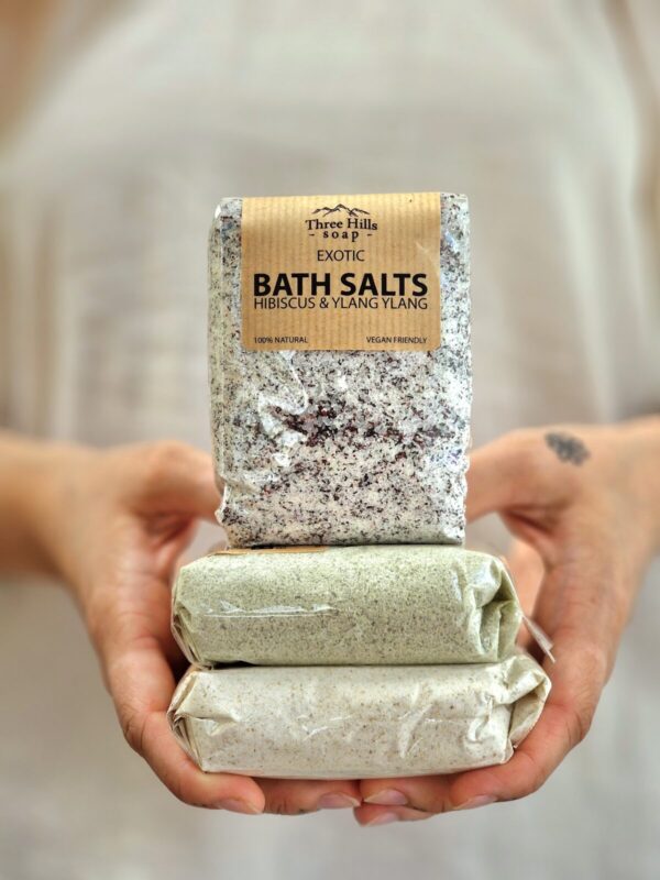 The image shows a person holding three packages of bath salts stacked on top of each other. The top package is labeled "Three Hills Soap Exotic Bath Salts Hibiscus & Ylang Ylang," and is described as 100% natural and vegan-friendly. The bath salts inside this package appear to have a mixture of white and purple granules. The other two packages below are wrapped in a plain material, one light green and the other off-white, but their contents and labels are not visible. The focus of the image is on the packages, with the background being softly blurred.