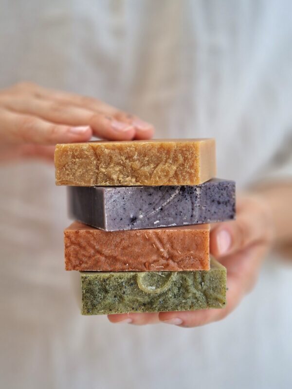 The image shows a person holding a stack of four handmade, natural soap bars, each in different colors and textures. The soap bars are rectangular and are stacked one on top of the other. The top bar is a light brown color, followed by a dark grayish-purple bar, an orange-brown bar, and finally, a green bar at the bottom. The texture of the soaps suggests they are handcrafted, with natural, rough edges and visible ingredients embedded within them. The person's other hand is gently touching the top soap bar. The background is softly blurred, keeping the focus on the vibrant colors and textures of the soap bars.