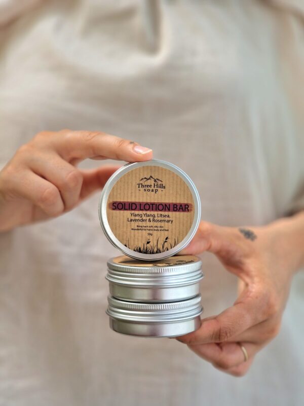 The image shows a person holding a stack of three small, round tins, with the top tin's lid open to reveal the product inside. The open tin is labeled "Three Hills Soap Solid Lotion Bar." The label indicates that the lotion bar contains Ylang Ylang, Litsea, Lavender, and Rosemary, with the tagline "Bring back soft, silky skin. Wonderful for hands, body, and feet." The tin is 50g in size. The other tins below are closed, and their labels are not visible. The background is softly blurred, emphasizing the focus on the lotion bar tins. The overall aesthetic is natural and minimalistic, with a neutral color palette.