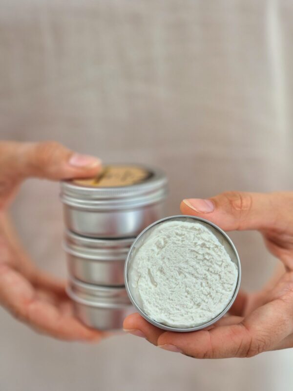 The image shows a person holding a small, round tin of natural deodorant cream. The tin in the right hand is open, revealing a white, creamy product inside, with a slightly textured surface. The person’s left hand is holding a stack of closed tins, similar in appearance to the open one. The focus is on the open tin, with the background softly blurred, creating a clean and minimalistic look. The overall presentation emphasizes the natural and handcrafted quality of the product.