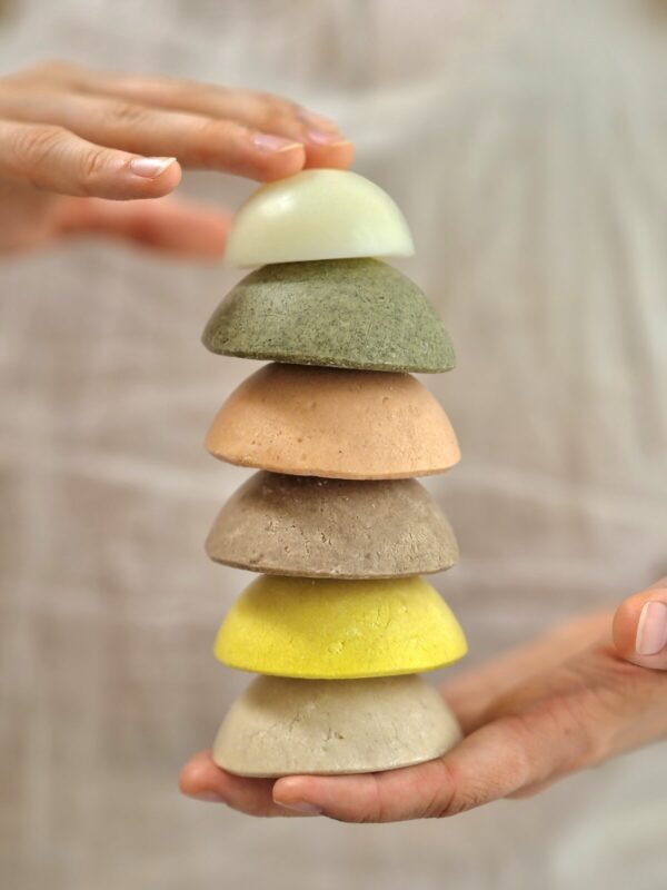 The image shows a person holding a stack of six dome-shaped shampoo and conditioner bars, each in different colors, suggesting different formulations or scents. The bars are neatly stacked on top of each other, creating a visually appealing tower. From top to bottom, the bars are colored white, green, peach, light brown, bright yellow, and beige. The person's left hand is gently touching the top bar, while the right hand is supporting the stack from the bottom. The background is softly blurred, keeping the focus on the colorful and varied textures of the shampoo and conditioner bars. The overall presentation emphasizes the natural and artisanal quality of the products.