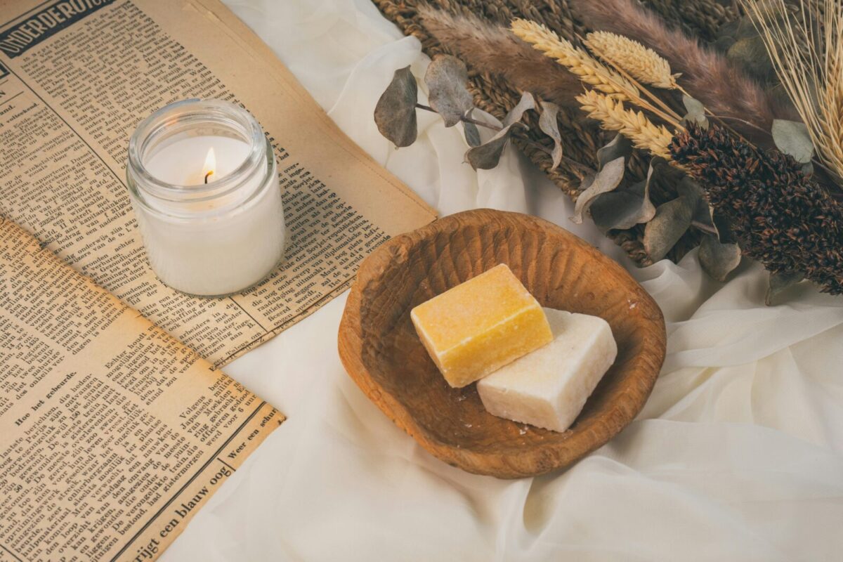 Artisan handmade soaps in a wooden dish surrounded by natural elements, with a lit candle and dried plants, creating a rustic and eco-friendly atmosphere.