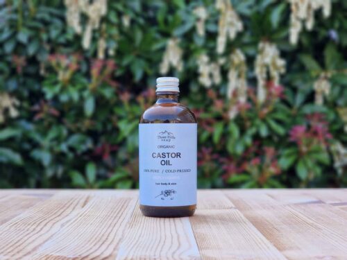 Three Hills Soap Organic Castor Oil in 100ml amber glass bottle with silver cap, displayed on outdoor wooden surface with green foliage background