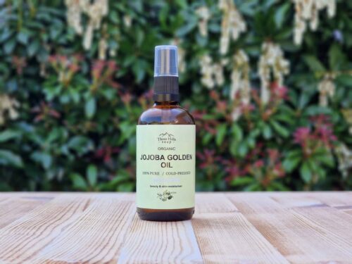 Three Hills Soap Organic Jojoba Golden Oil in 100ml amber glass bottle with pump, displayed outdoors on wood with blurred greenery